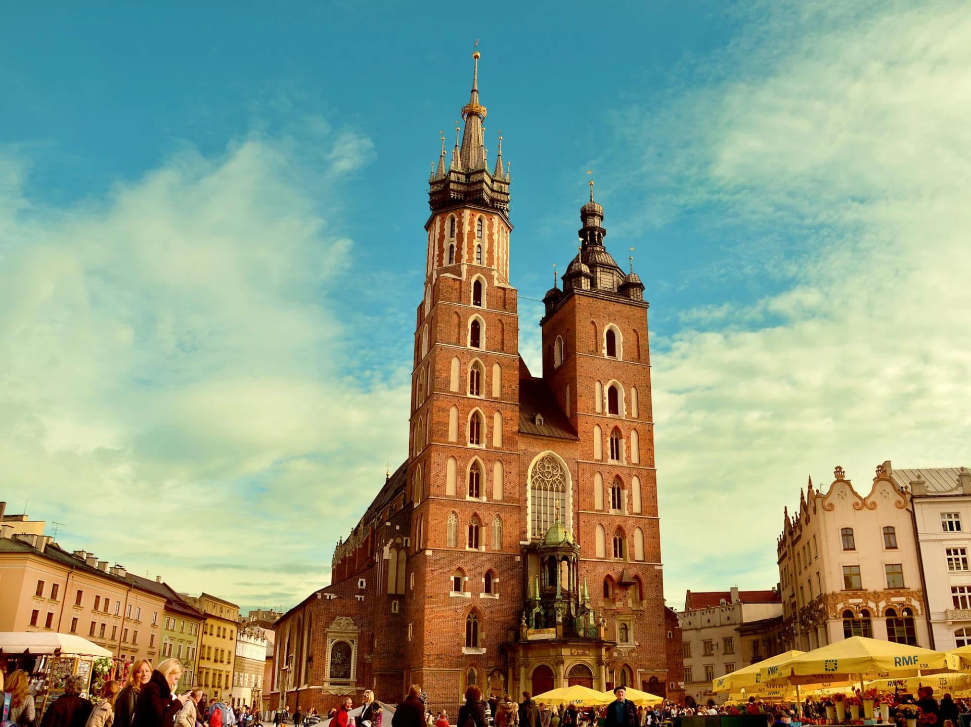 The image shows a historic brick church with two tall towers, surrounded by people and outdoor market stalls under yellow umbrellas.