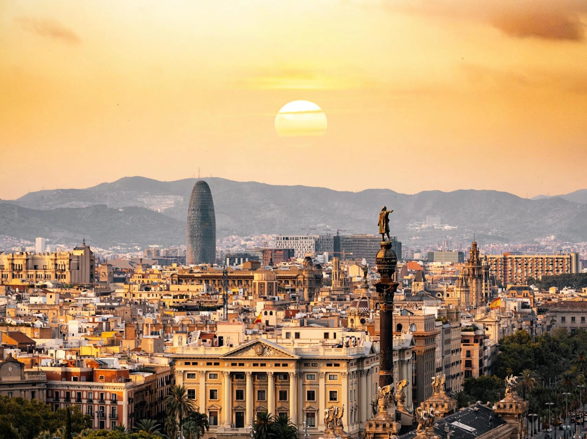 Sunset over Barcelona skyline with prominent buildings and statues in the foreground.