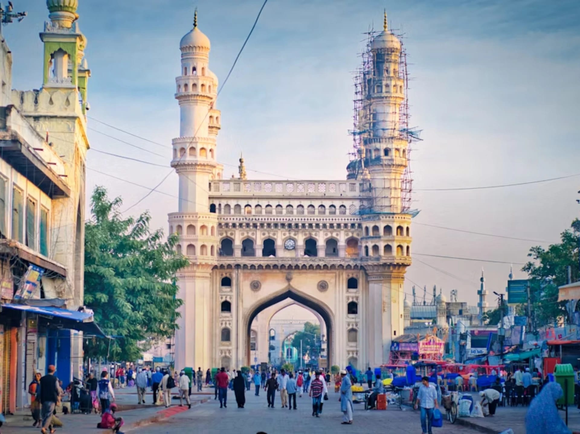A busy street scene in front of a large, ornate gateway with two minarets, some under construction scaffolding, and people walking and shopping.