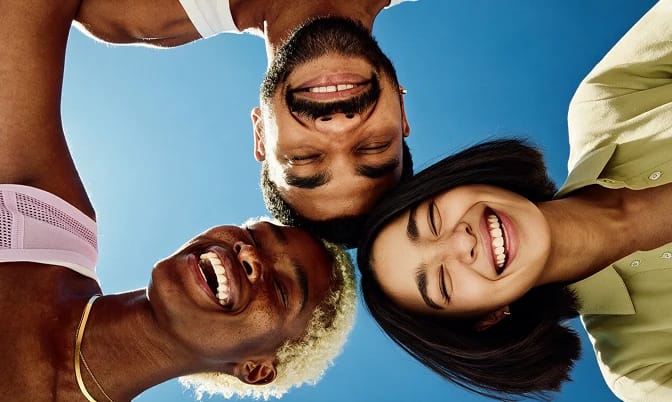 Four friends smile as they lean over the camera outdoors against a blue sky.