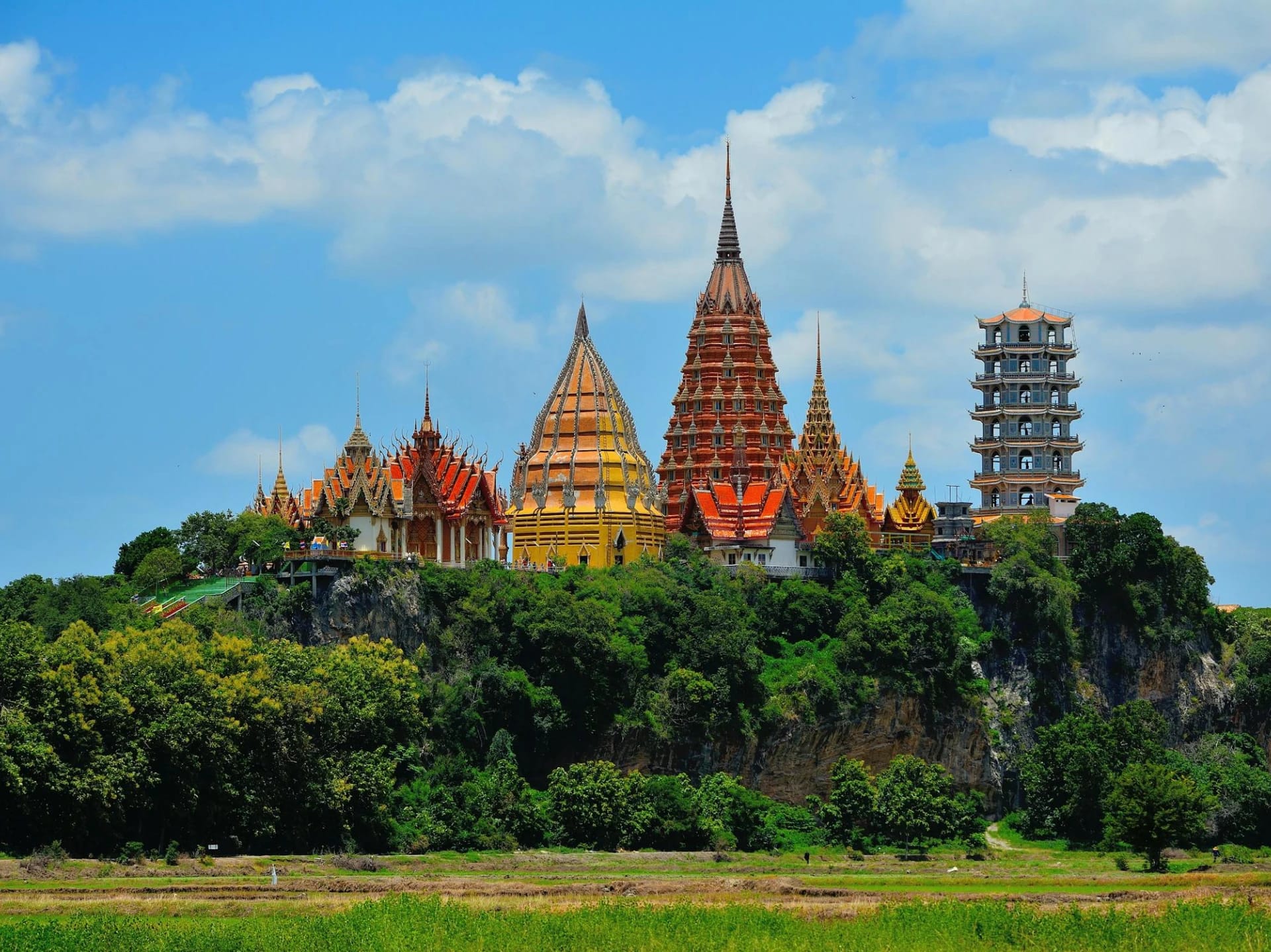 A colorful temple complex on a hilltop surrounded by greenery under a blue sky with clouds.