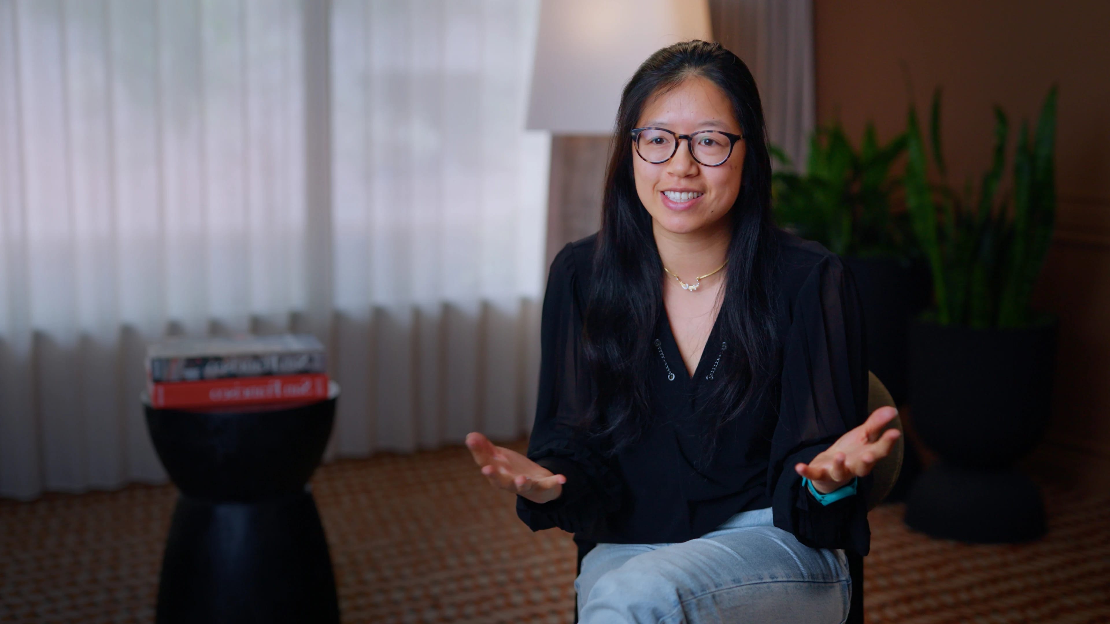 Woman wearing glasses speaks to the camera and gestures in a home setting.