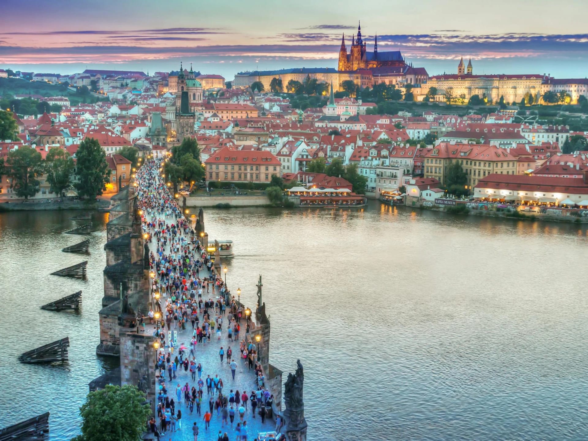 A crowded bridge over a river with a historic cityscape and castle in the background at sunset.